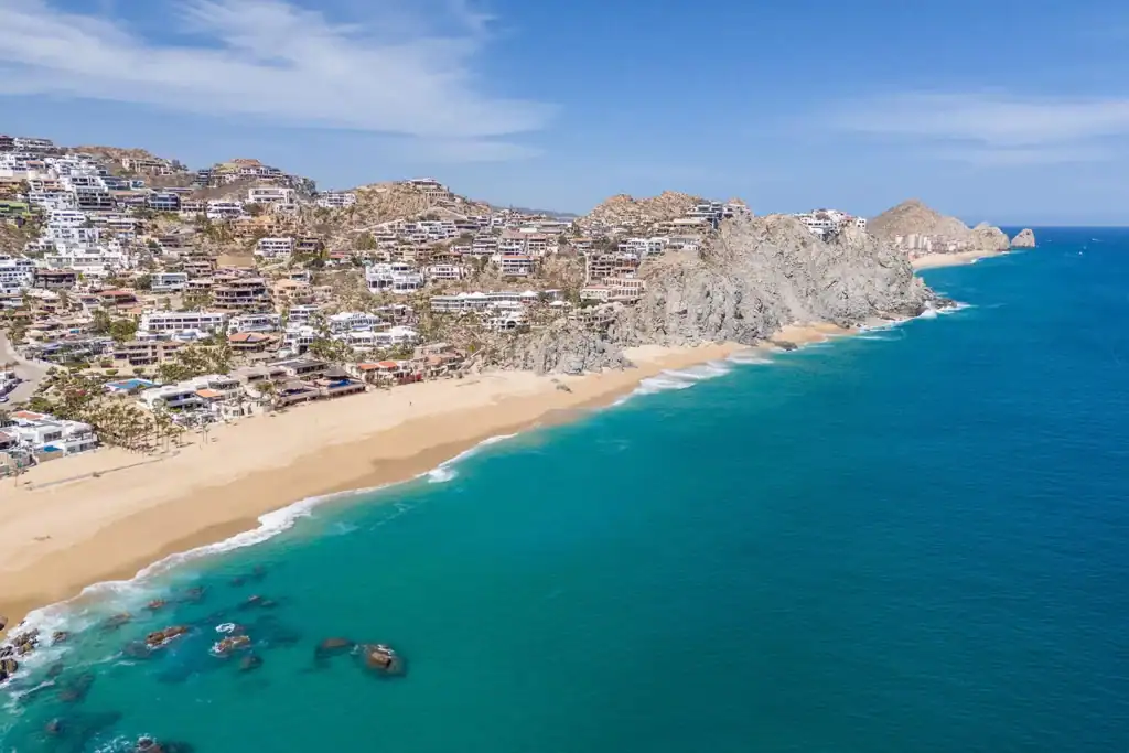 Aerial view of Pedregal de Cabo San Lucas with hillside luxury homes cascading toward a golden sand beach, granite cliffs, and turquoise Pacific Ocean waters with Land's End visible in the distance
