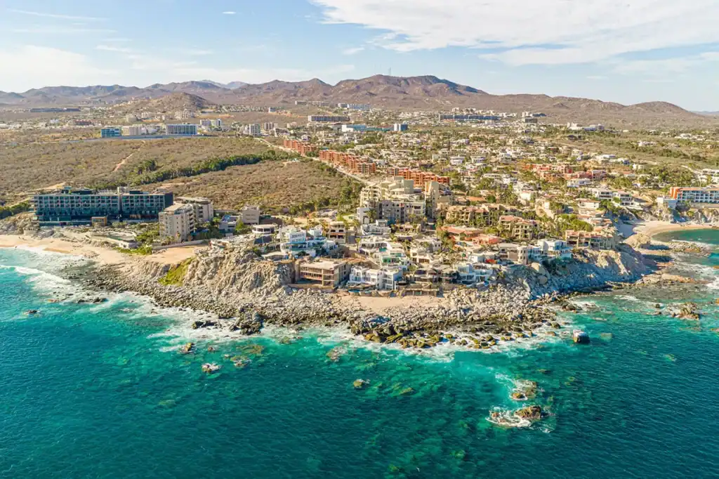 Aerial view of a rocky coastline with turquoise sea and a dense seaside town beyond, hills in the distance.