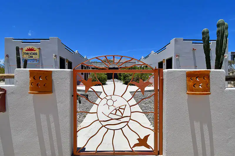Orange sun-themed metal gate at the entrance of a white stucco inn, with desert gravel and tall cacti against a bright blue sky.