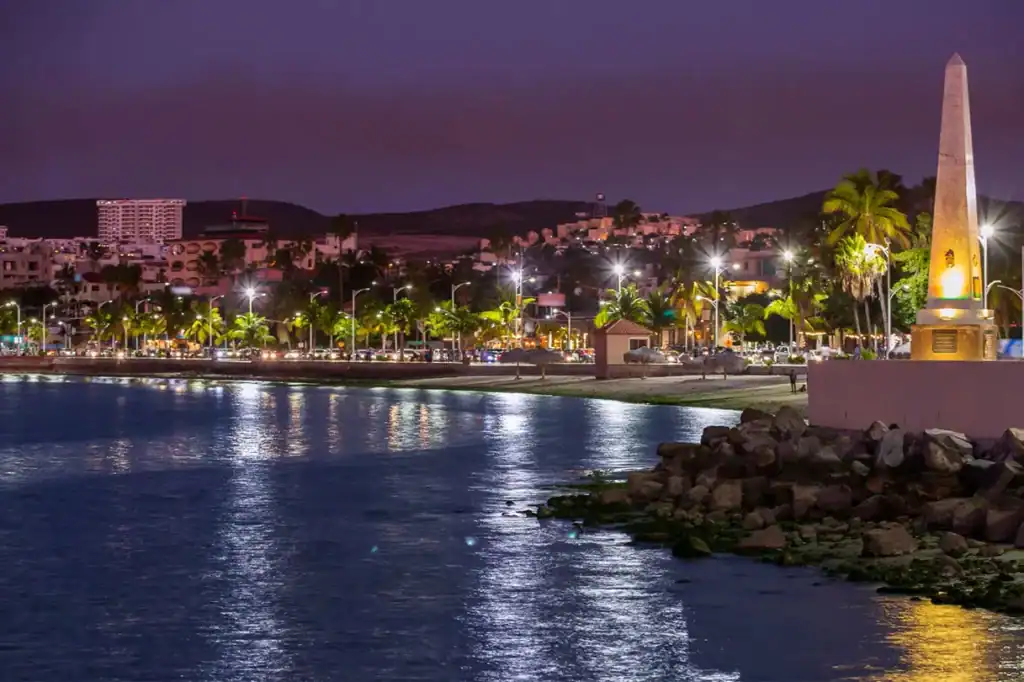 Night view of the La Paz Malecón waterfront promenade with illuminated palm trees, obelisk monument, and city lights reflecting on the Sea of Cortez, Baja California Sur, Mexico.