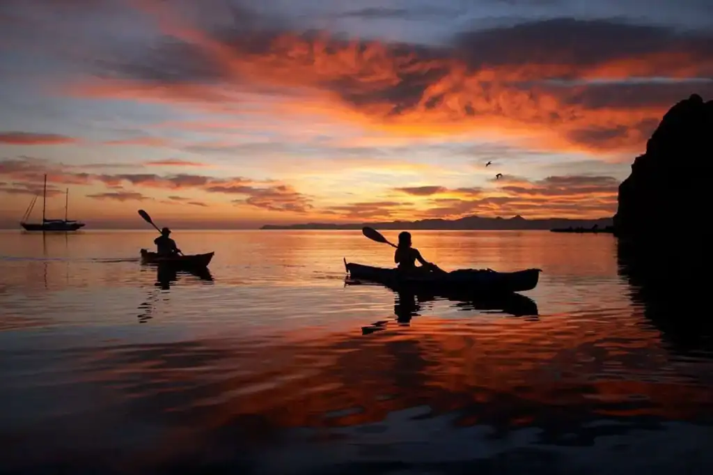 Two kayakers paddling on the Sea of Cortez at sunset near Loreto with a sailboat and island silhouette in the background, Baja California Sur, Mexico.