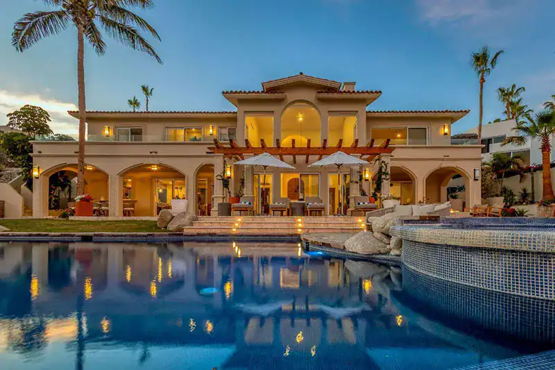 Luxurious two-story villa with arches, palm trees, and a reflecting pool at dusk.