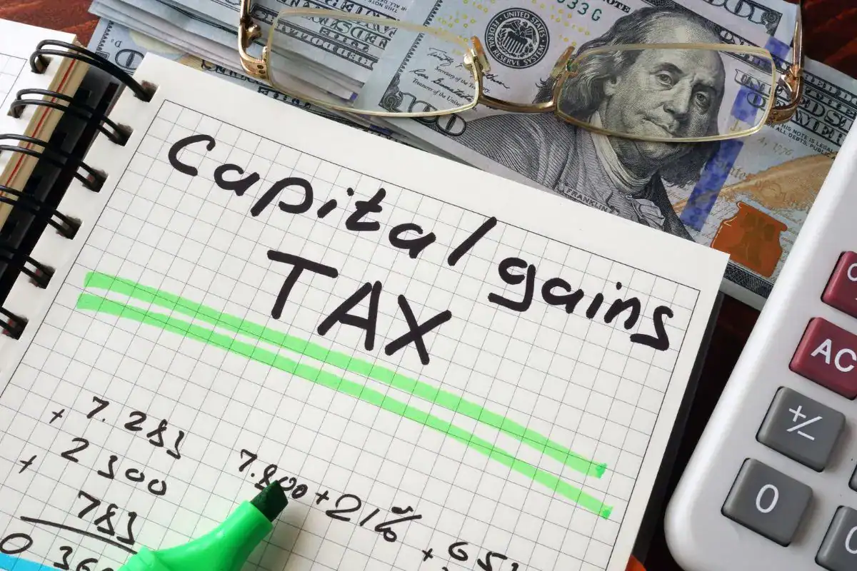 Notebook with capital gains tax written and underlined, beside US dollar bills, reading glasses and a calculator